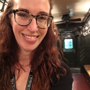 Leigh poses with long hair and glasses in front of dark background