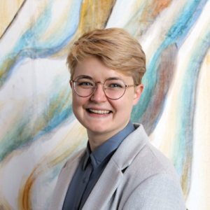 a white person with blonde hair, glasses, and a light blue coat smiles directly at the camera in front of abstract art