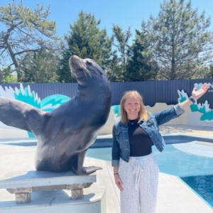 A white woman in a blue coat and white pants poses with an arm up next to a seal